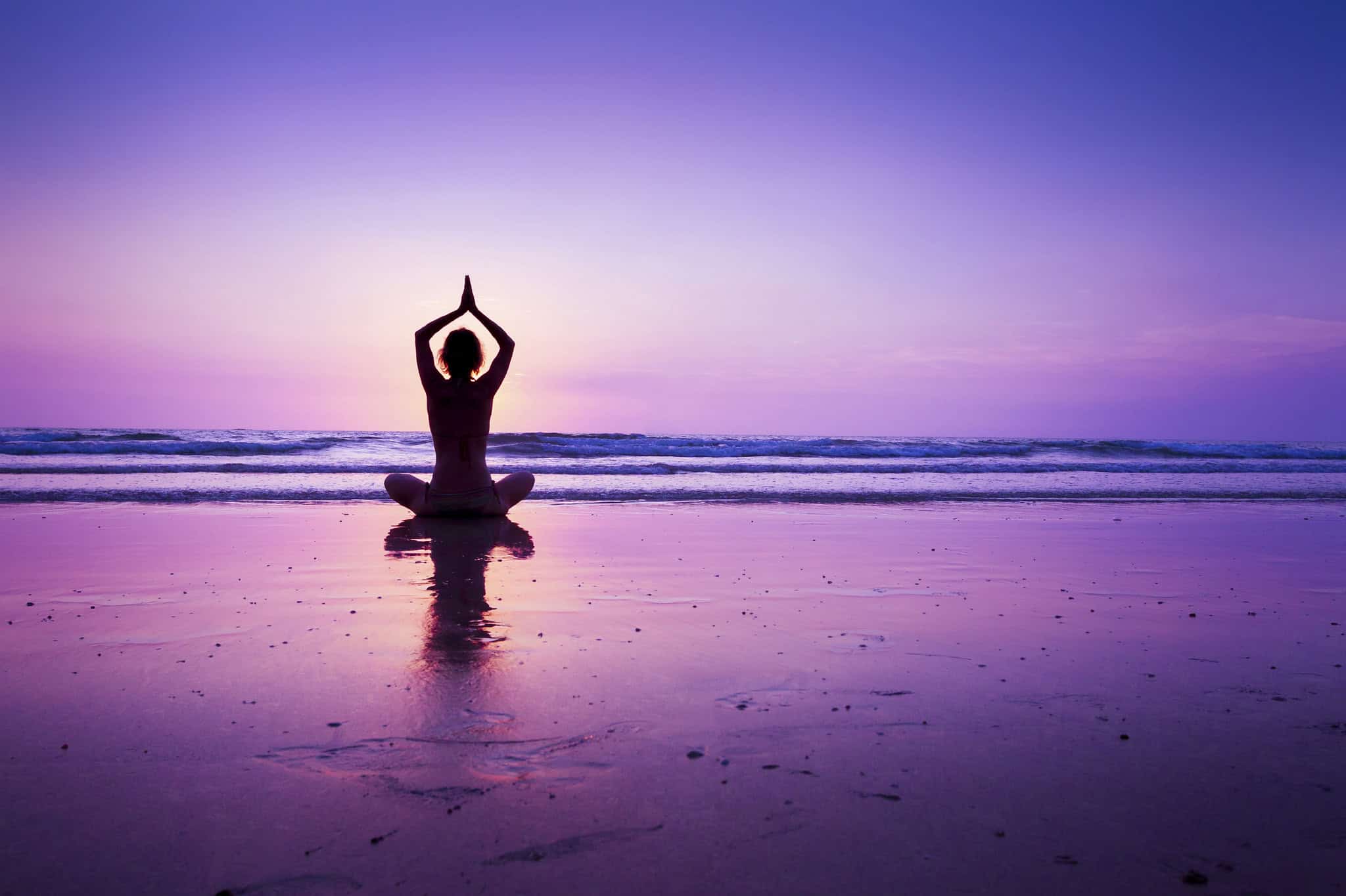 a seated woman doing yoga on a beach at sunrise with her arms raised above her head.