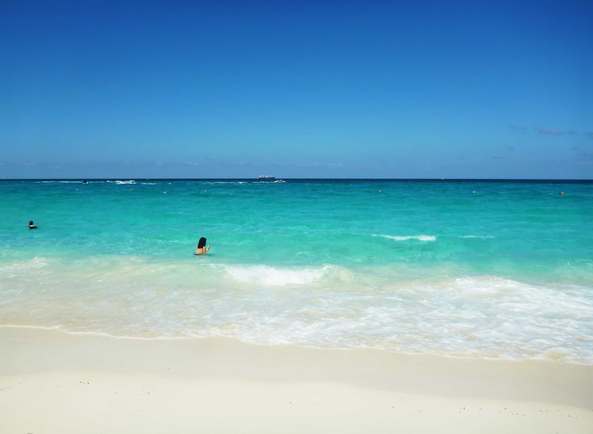 a woman swimming in turquoise water looking out toward the horizon with a white sand beach in the foreground and blue sky above