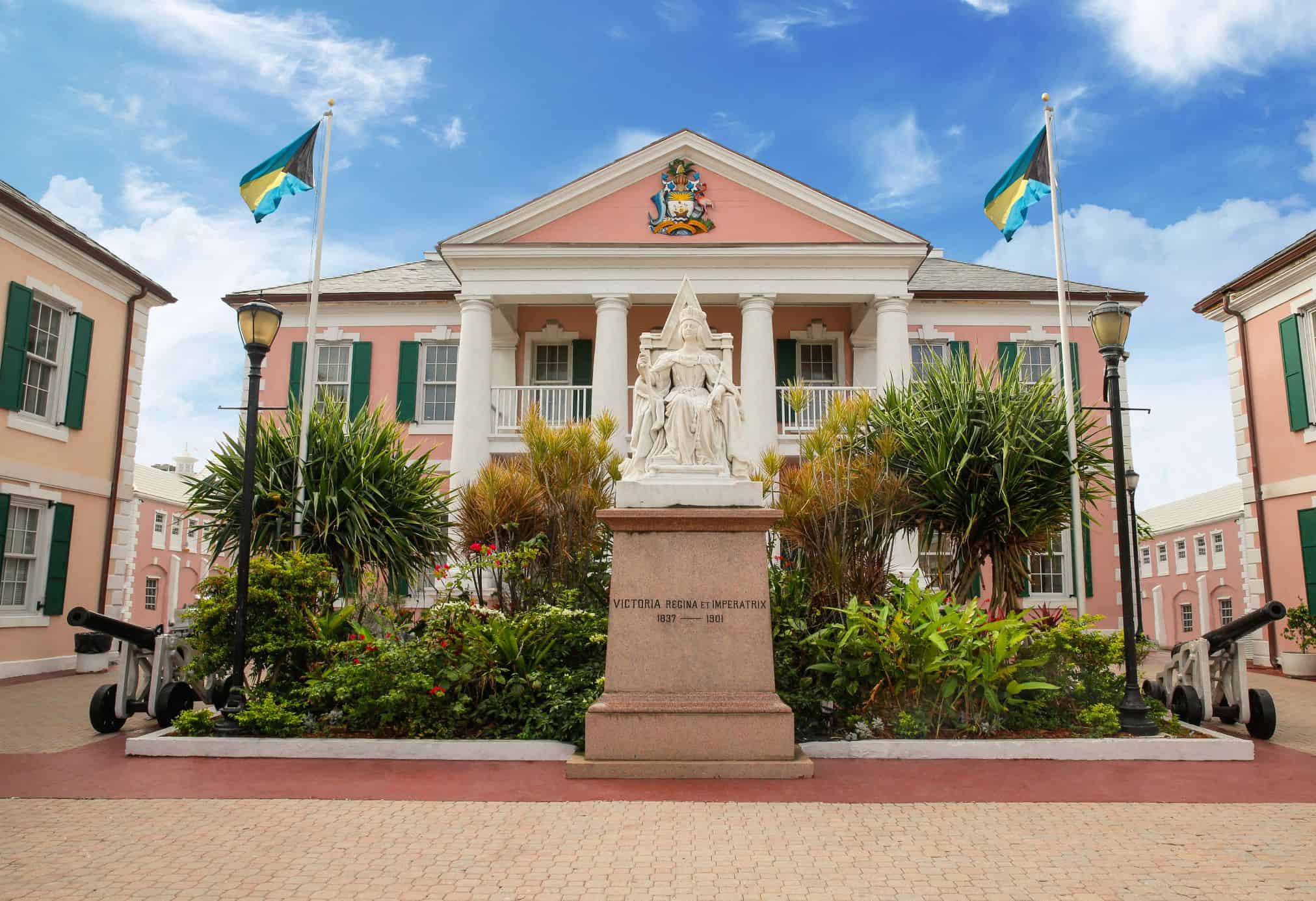 Pink building with a statue in front and two flags of the Bahamas floating in the breeze