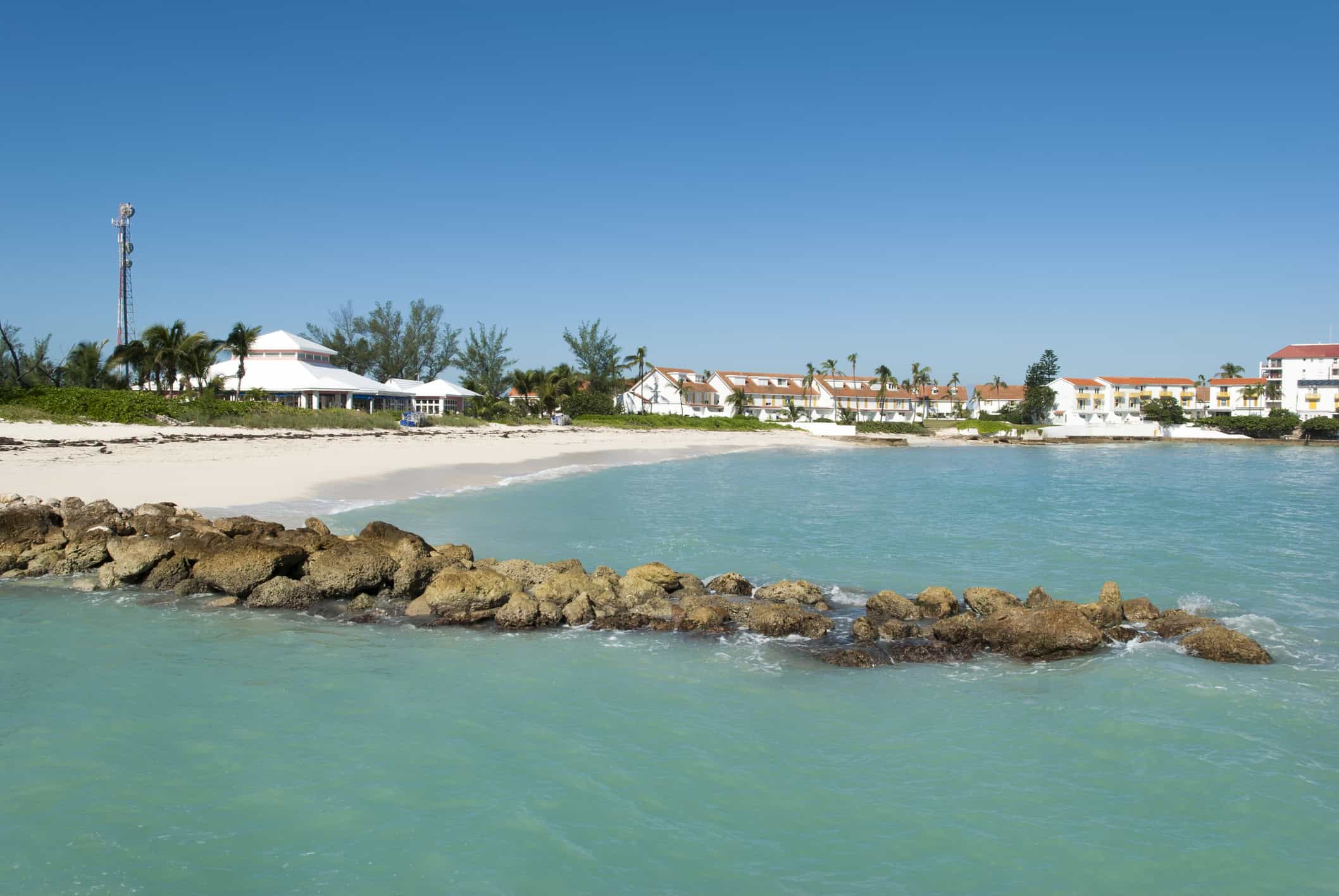 turquoise water with stone breakwater in foreground, a sand beach behind it edged by low white buildings